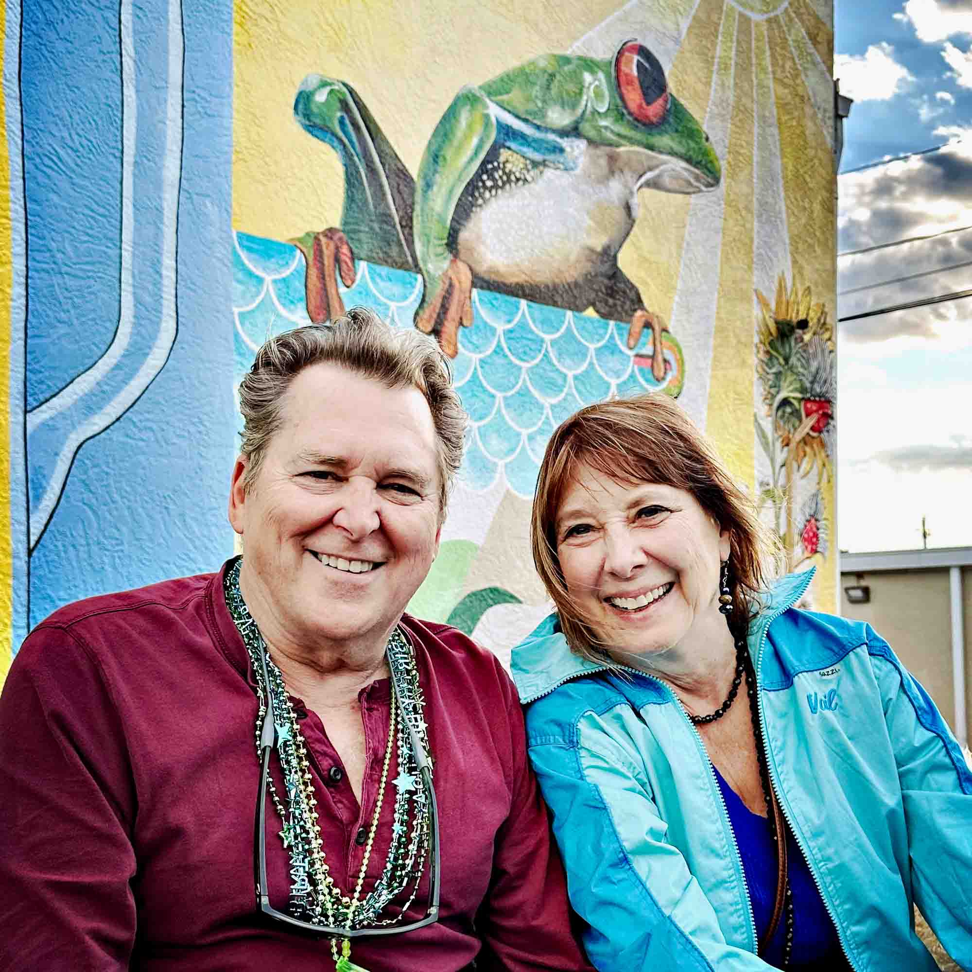 Smiling couple sits together in front of a colorful mural featuring a frog, wearing festive beads and enjoying a cheerful day.